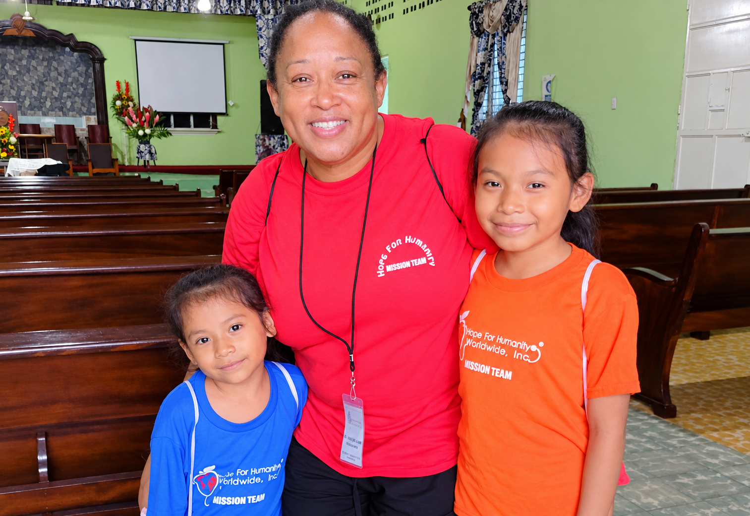 Hope for Humanity volunteer in red mission team shirt hugging two children in HHW shirts inside church hall