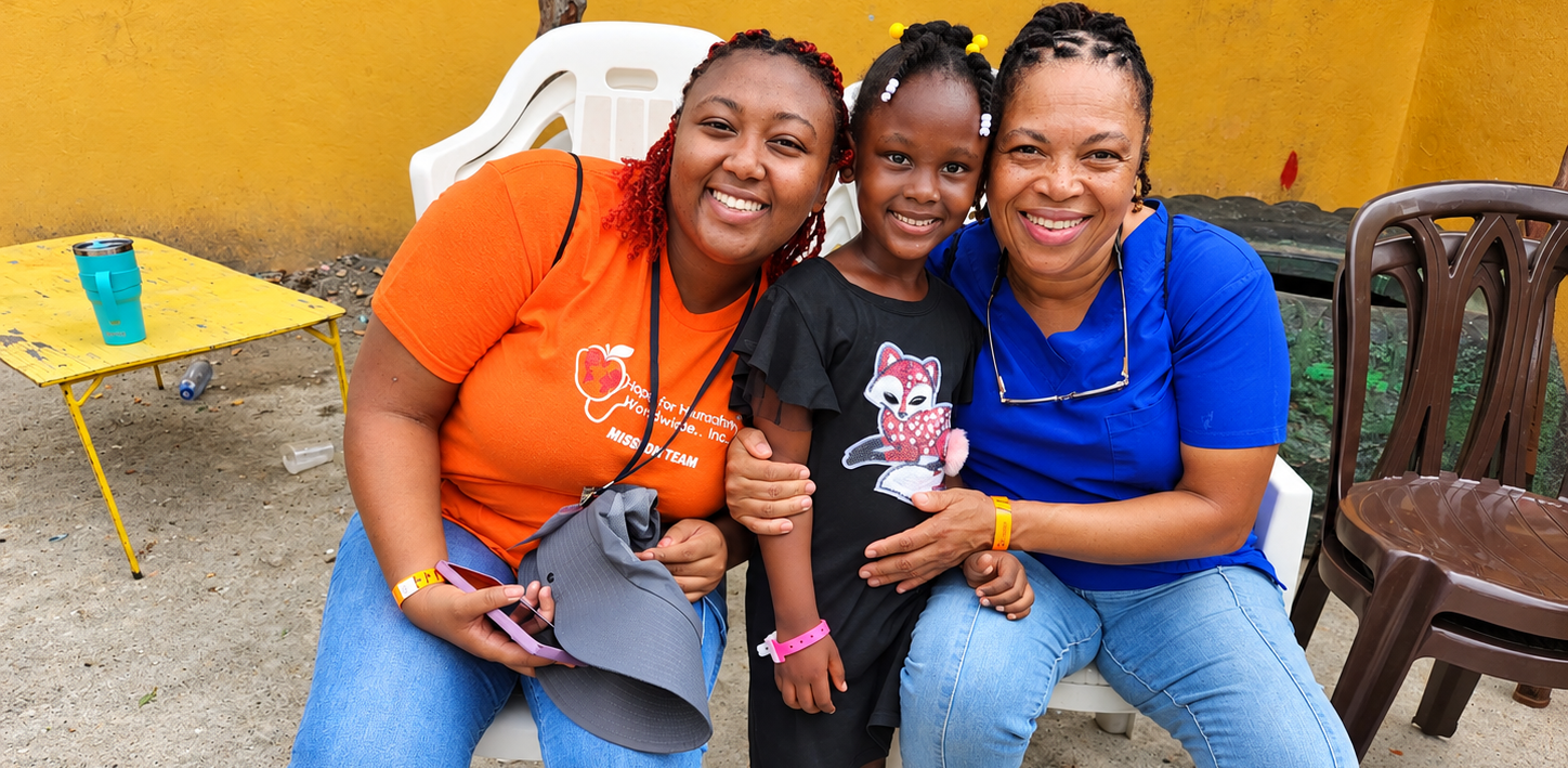 Two Hope for Humanity volunteers in orange and blue shirts hugging a young girl outdoors against yellow wall