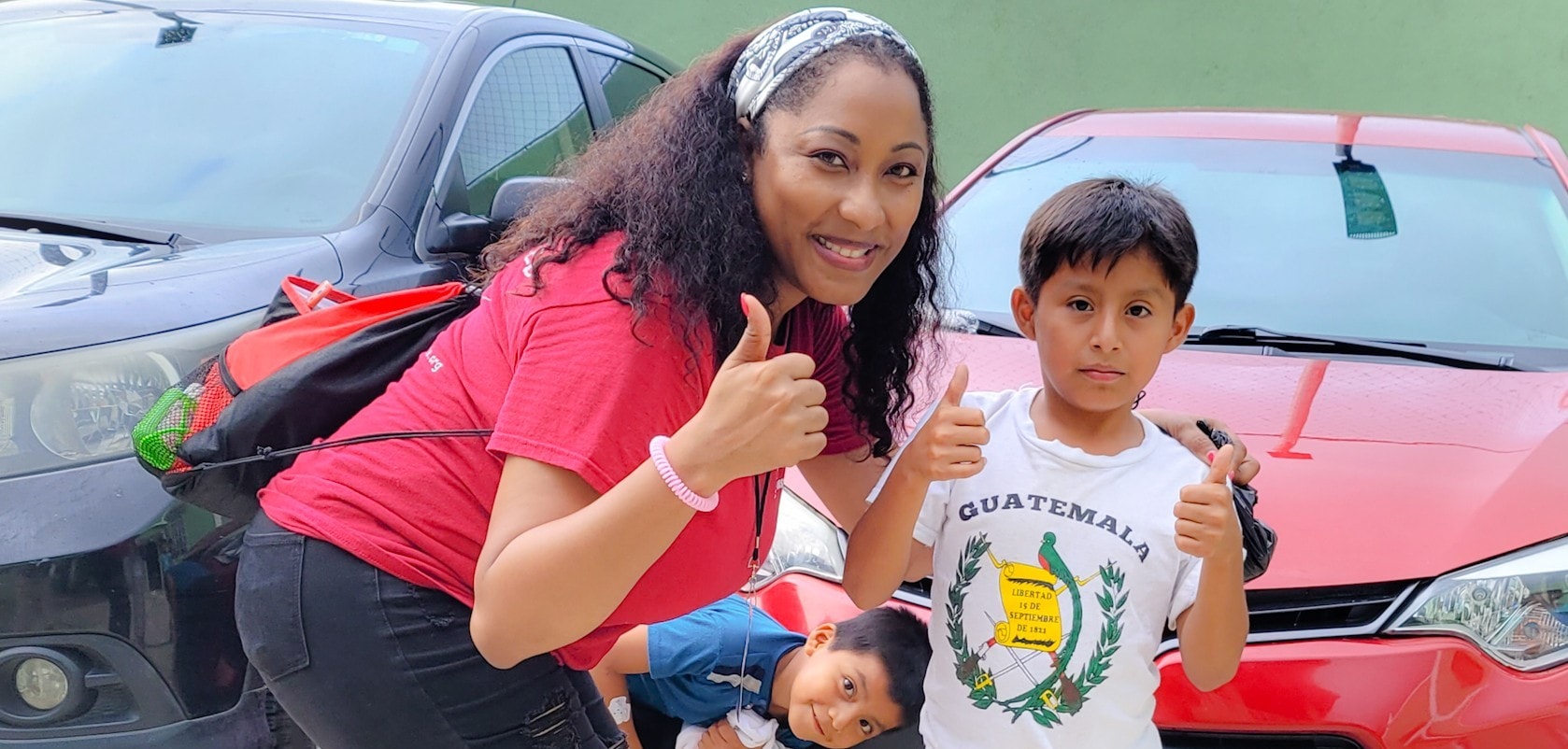 Hope for Humanity volunteer in red shirt giving thumbs up with two Guatemalan children in parking lot