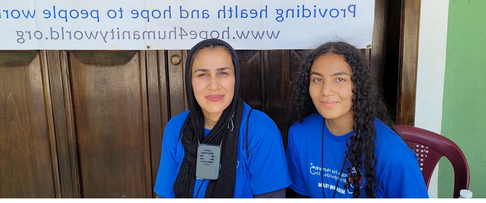 Two Hope for Humanity volunteers in blue mission team shirts seated in front of HHW banner at community clinic