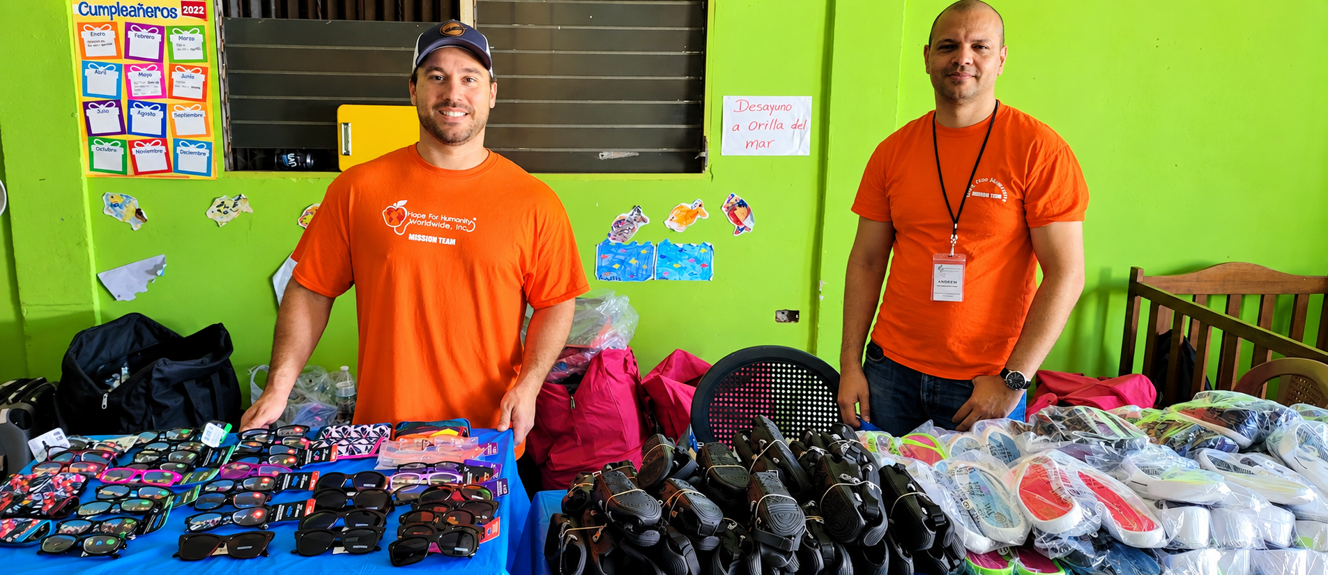 Two Hope for Humanity male volunteers in orange mission team shirts standing behind table of sunglasses and sandals donations