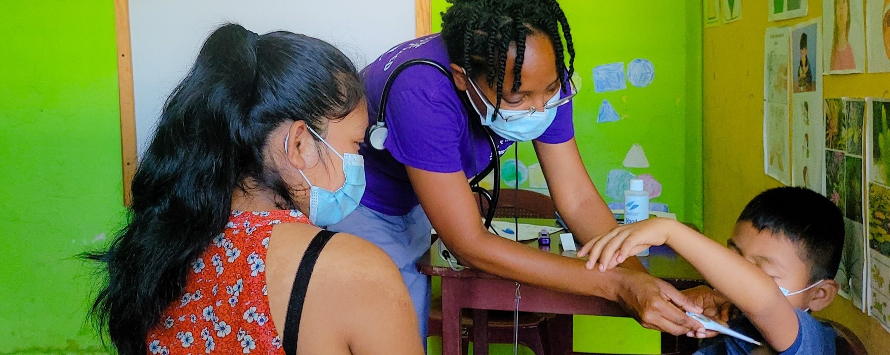 Hope for Humanity volunteer in purple shirt with stethoscope examining young child with mother present in green classroom