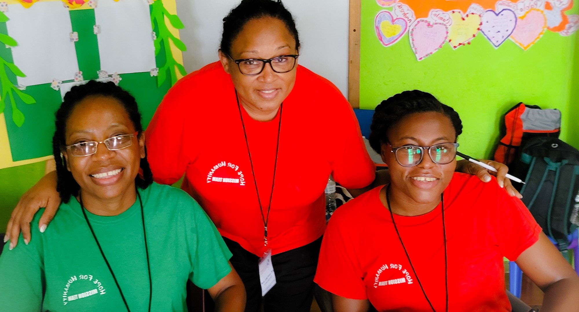 Three Hope for Humanity mission team members in red and green shirts smiling together inside colorful classroom