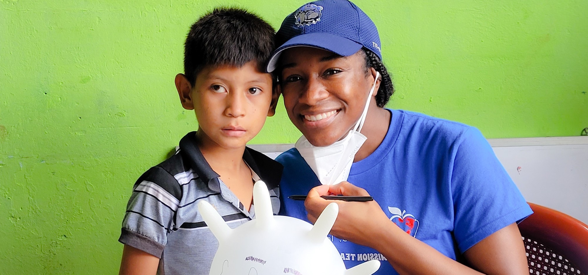 Hope for Humanity volunteer in blue mission team shirt smiling with young boy holding craft project against green wall