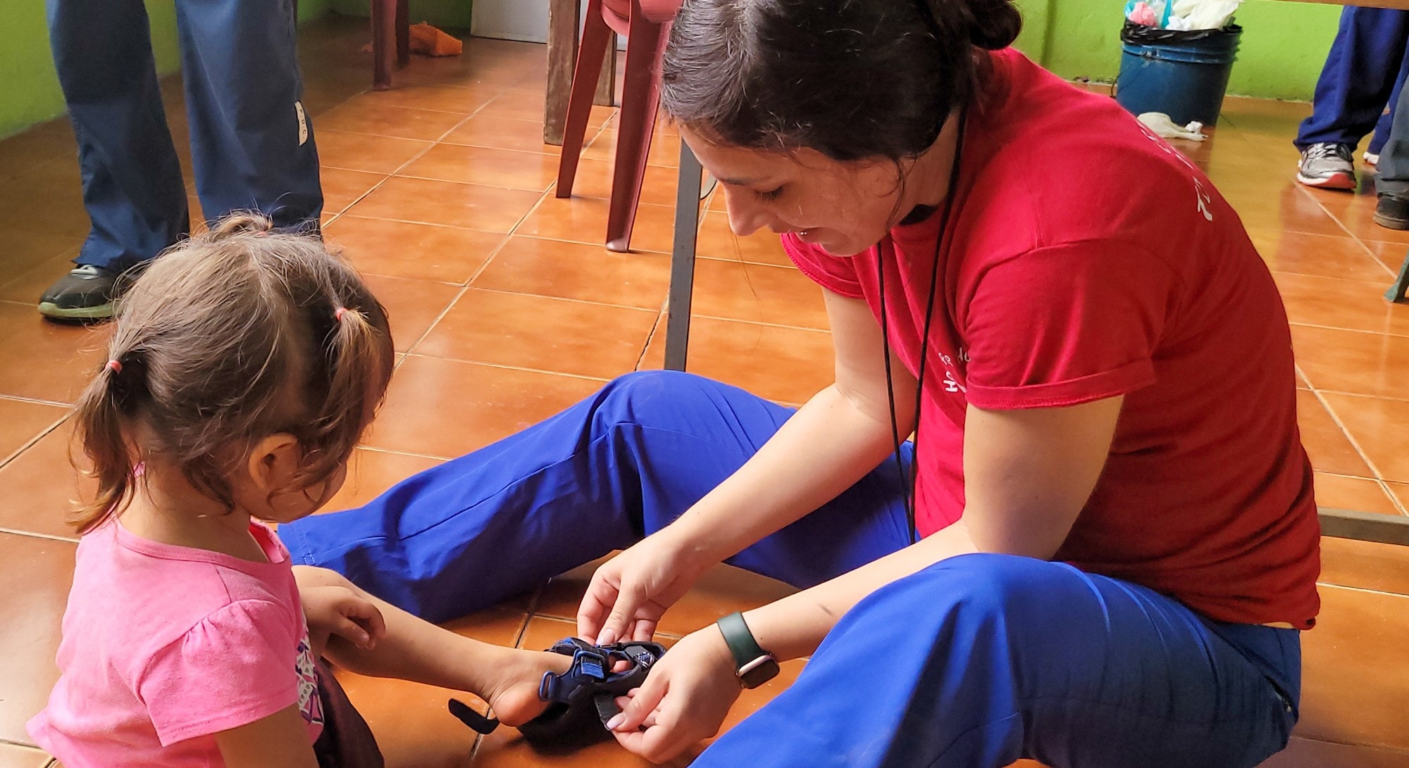 Hope for Humanity volunteer in red shirt sitting on floor helping toddler girl with shoes at indoor community clinic